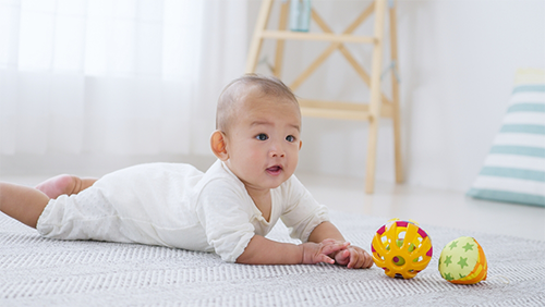 A 6-month-old Asian baby on a floor mat at home trying to reach for a toy ball in front of him