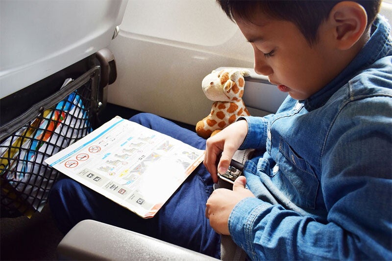 A boy putting on seatbelt in plane while reading flight instructions and with a stuffed giraffe. 