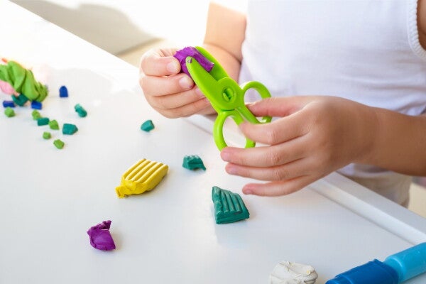 A cropped photo of a little girl’s hands holding green safety scissors in one hand and clay in the other.