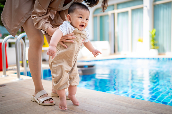 A cropped photo of a mom holding her 1-year-old son outdoors near a pool, helping him take his first few steps.