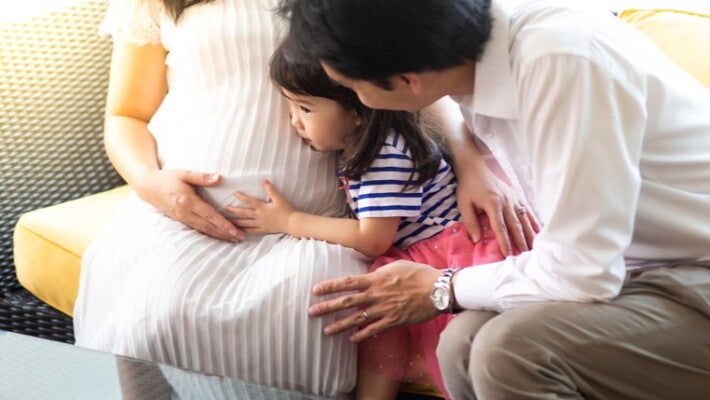A little girl has her ear to her mom’s pregnant belly while the father looks on.