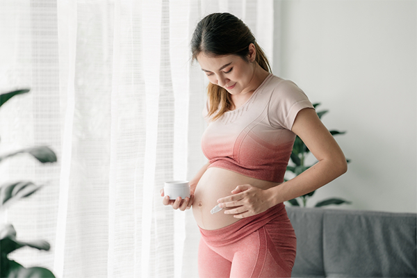A pregnant Asian woman applying cream on her belly while standing in her living room.