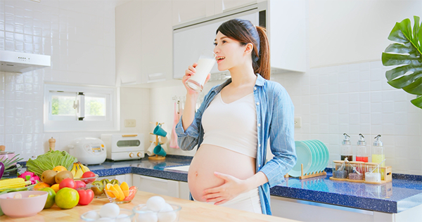 : A pregnant Asian woman drinking a glass of milk, with vegetables and fruits on the kitchen island in front of her