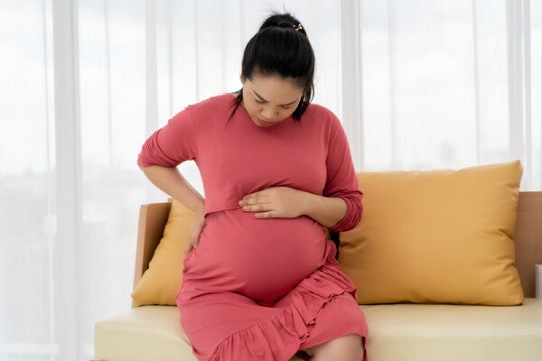A pregnant Asian woman sitting down in bed, with her hand below her chest, feeling discomfort.