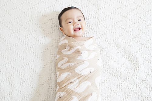 A smiling Asian baby boy swaddled in a printed wrap in bed