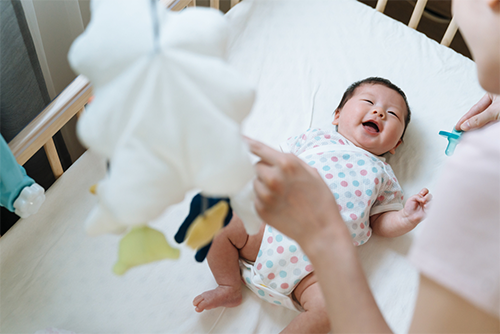 A smiling Asian baby in a crib looking up at her mom holding a pacifier and pointing to the mobile