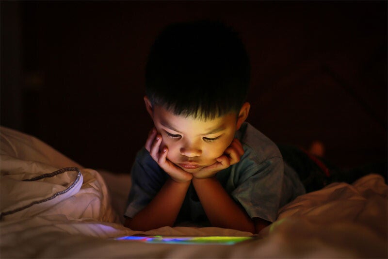 A young Asian boy watching a show on a tablet with his chin on hands, alone during bedtime, like a gadget addict