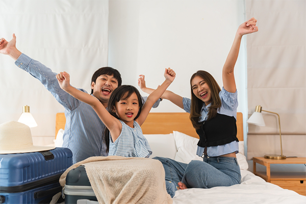 A young Asian couple and their preschooler daughter cheer while on the bed with their luggage.
