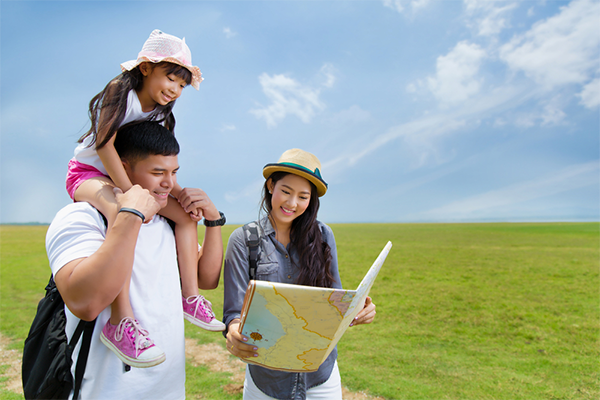 A young Asian dad carrying his little girl on his shoulder while the mom holds a map for them all to look at outdoors.