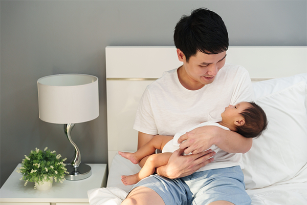 A young Asian dad looks at his baby sleeping in his arms.
