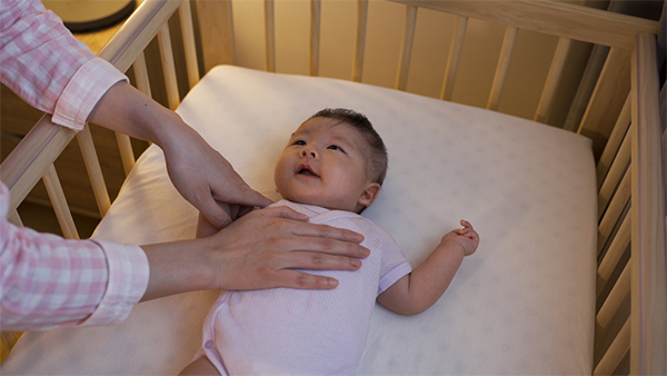 A young Asian mom placing her baby awake in a crib at night.