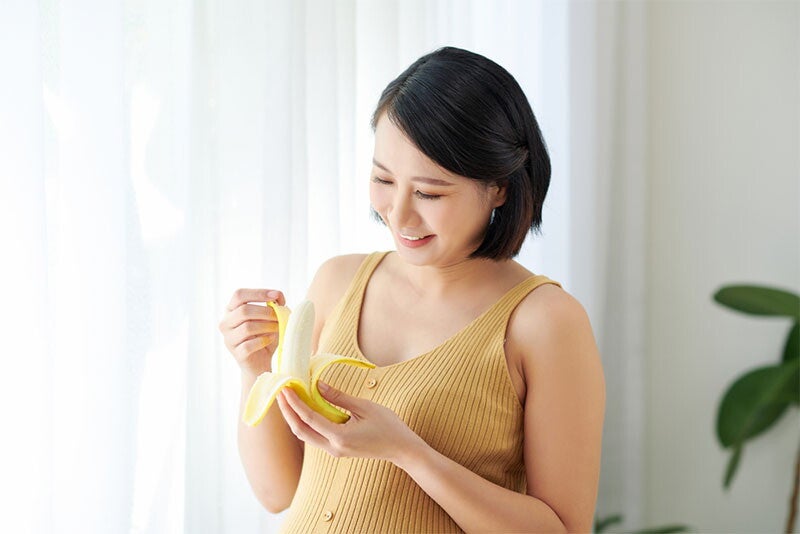 A young Asian pregnant woman is smiling and eating a banana.