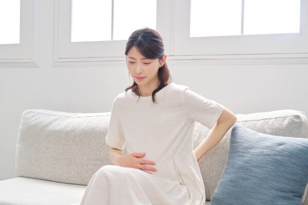 A young Asian woman holding her baby bump and her back while seated on the sofa at home.