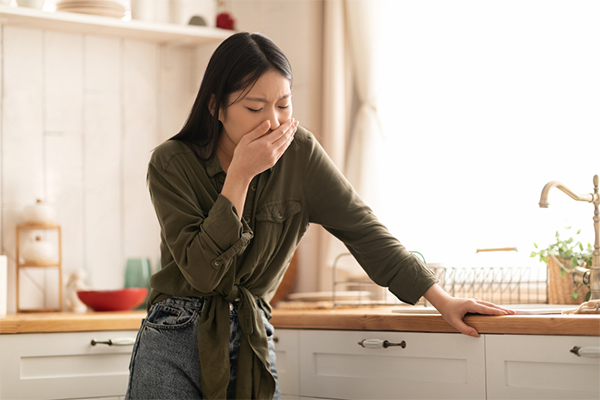 A young Asian woman in the kitchen, covering her hand with her mouth and feeling nauseated.