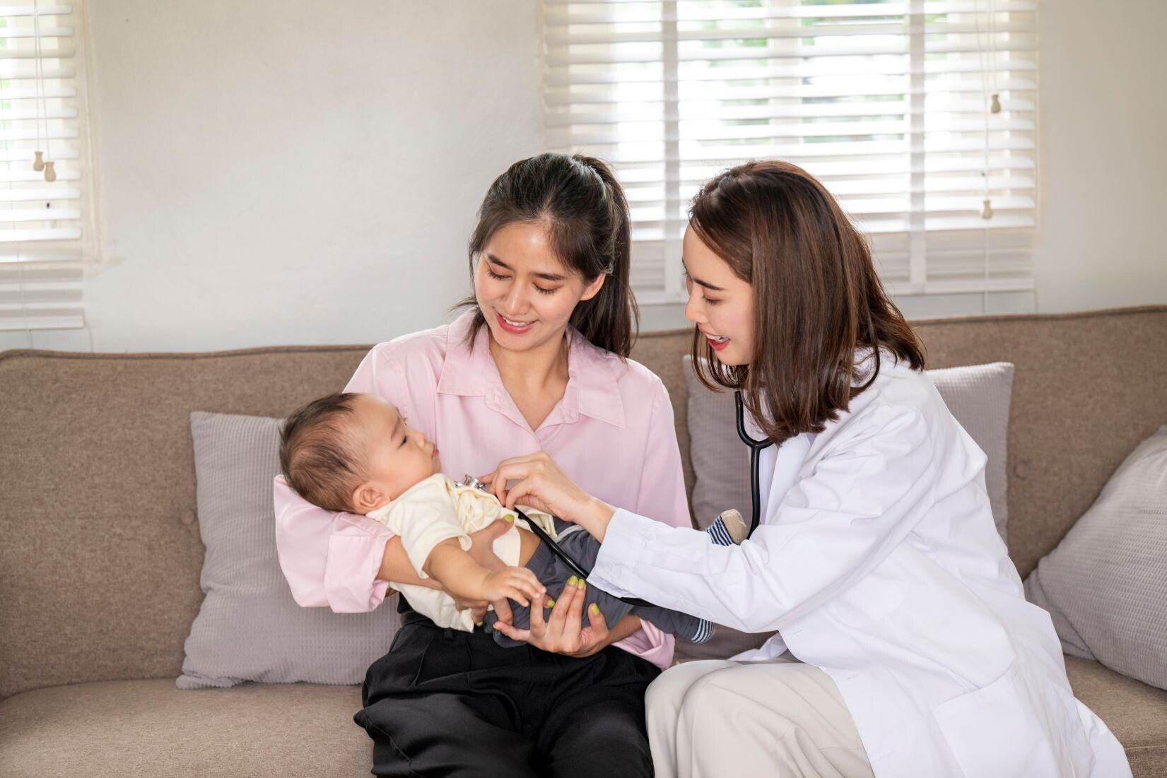 A young Asian woman is holding a baby on a couch while a doctor examines the baby.