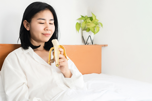 A young Asian woman lying in bed and eating a banana to help ease her nausea at 7 weeks pregnant