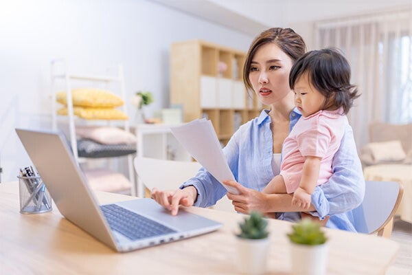 A young Asian woman seated at her desk, carrying her baby while holding documents in front of her laptop.