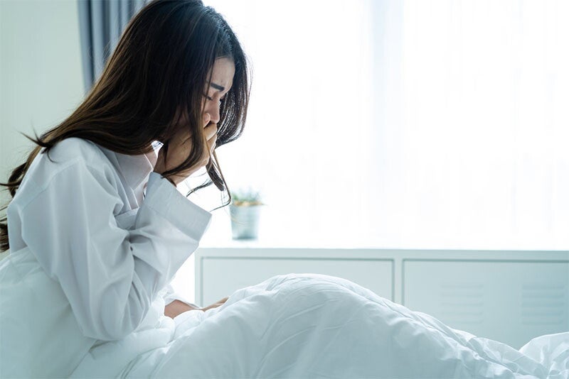 A young Asian woman sitting up in bed and covering her mouth with her hands, suffering from morning sickness