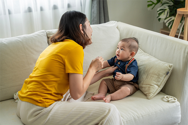 A young Asian woman tries to talk to her baby as they sit on a couch at home.