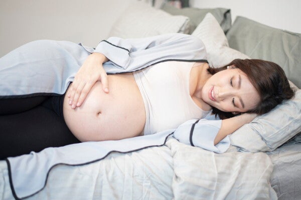 A young pregnant Asian woman lying on her sidein bed while looking at her baby bump.
