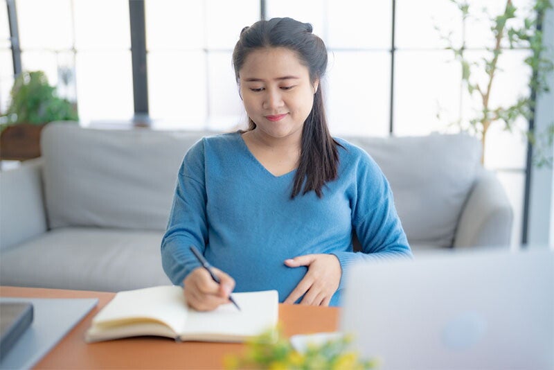 : A young pregnant Asian woman on her desk at home, making a list of things to prepare for delivery