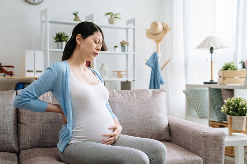 A young pregnant Asian woman on the sofa at home, trying to stretch her lower back