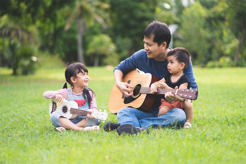 An Asian father and daughter playing guitar and ukulele together while the little baby boy is sitting with them.