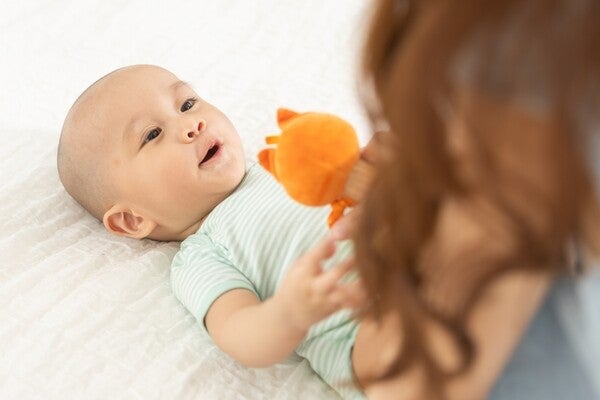 Asian baby looking at a bright orange toy his mom is holding for him while lying in bed.