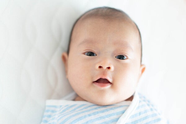 Asian infant lying on a white bed and looking at the camera with a neutral expression for a baby picture.