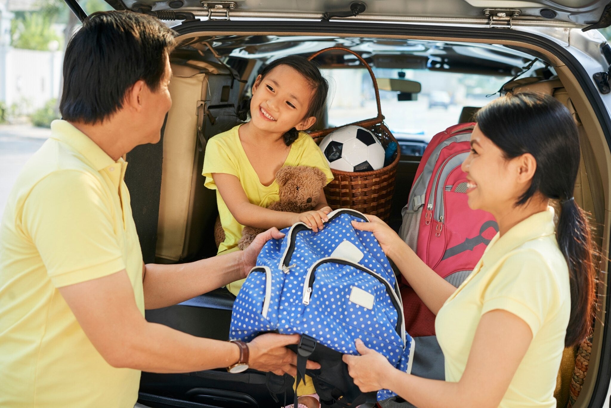An Asian couple and their little girl packing the trunk of their car for a family road trip.
