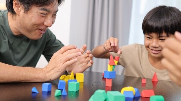 A smiling Asian father and son enjoy building structures with colorful geometric blocks indoors.