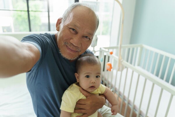 Asian grandparent with grandchild take a selfie with the crib behind him.