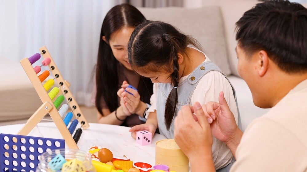 Asian parents guide their little girl who’s doing math for kids using a colorful abacus and dice.