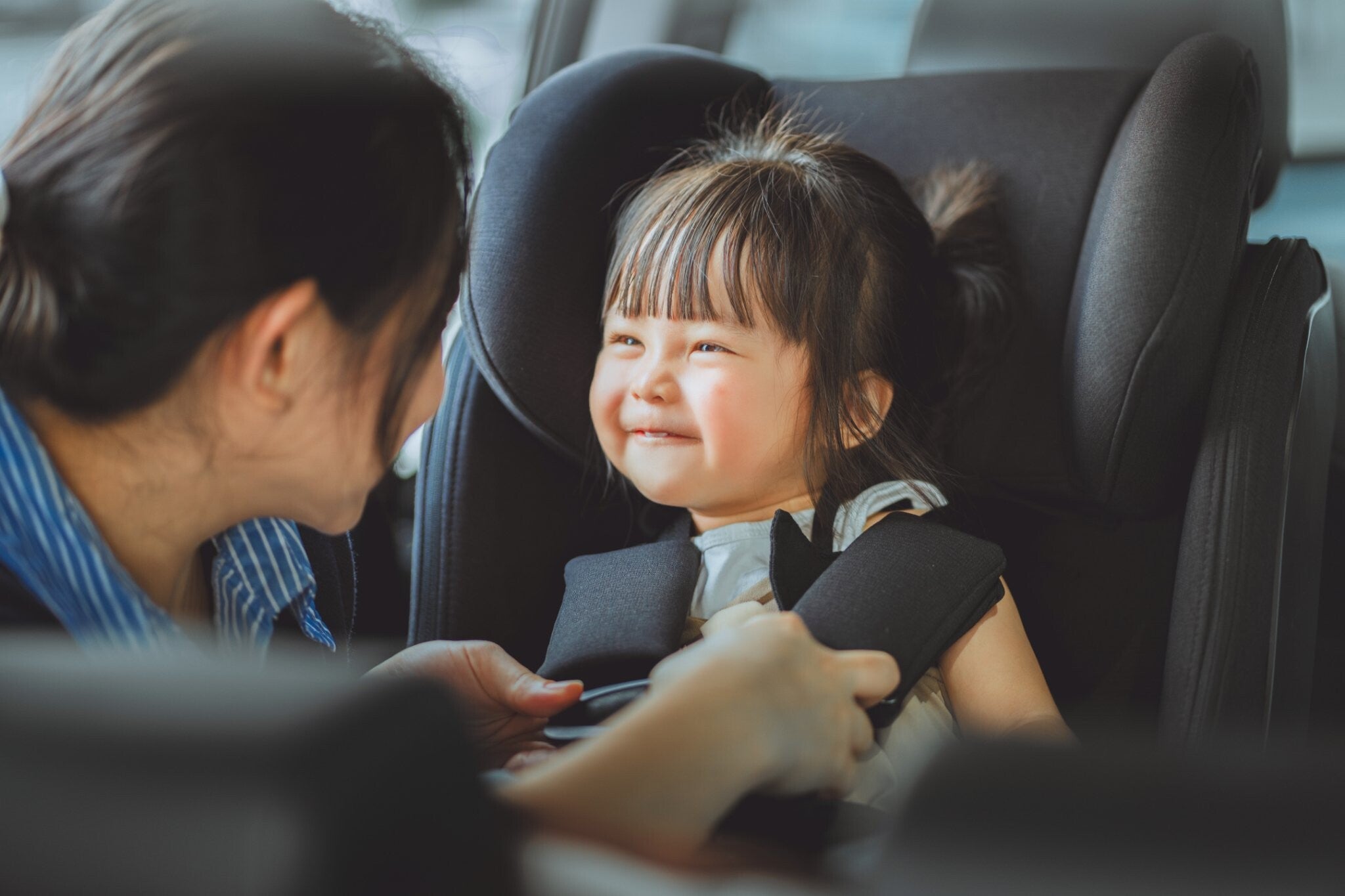Asian mom carefully buckles her smiling toddler daughter into a secure car seat before a road trip.