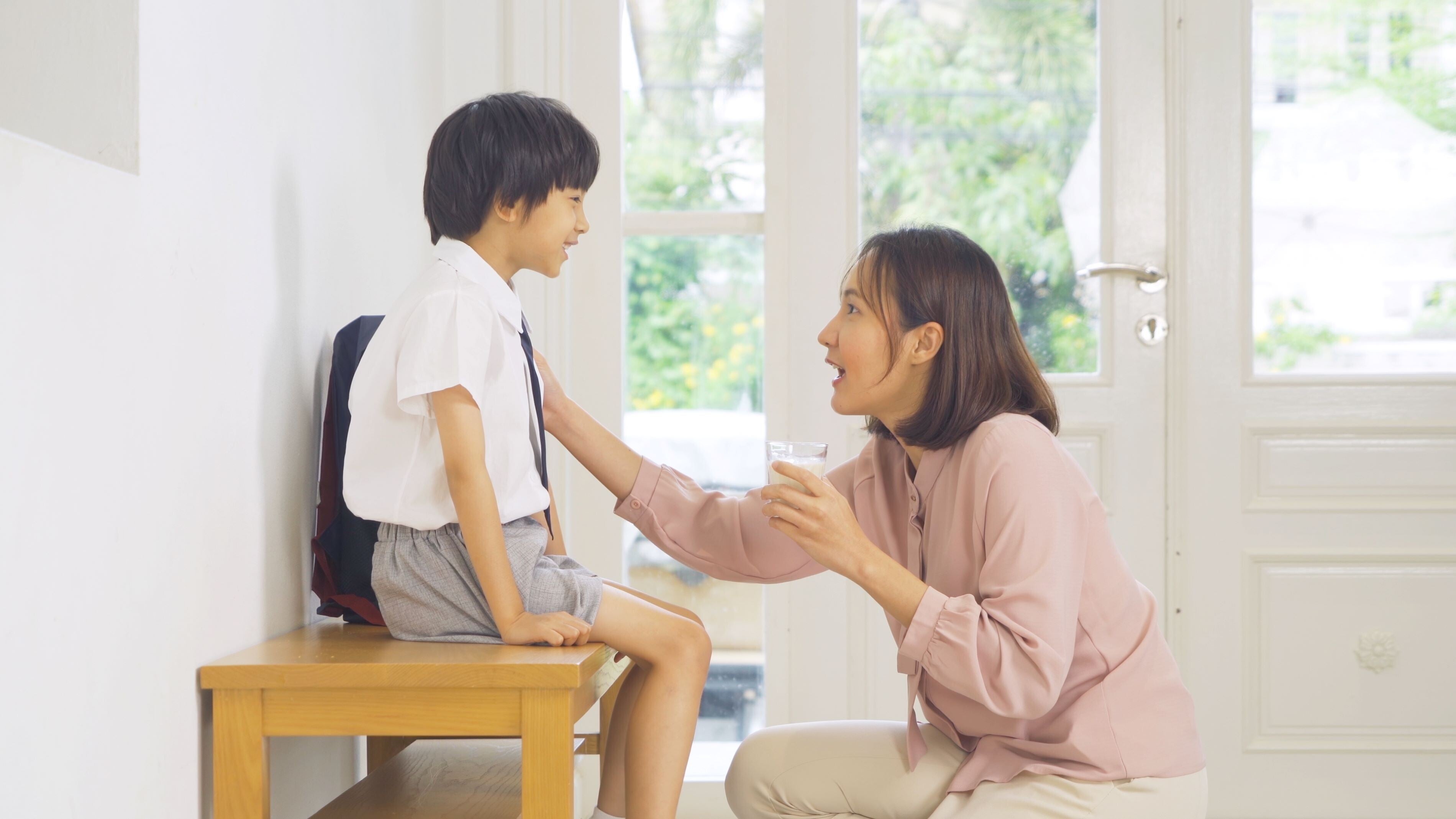 Asian mom giving a glass of fortified growing-up milk to her school-aged son in a uniform.