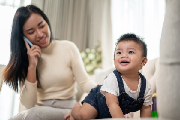 Asian mom on the phone in the background while her little son is smiling and crawling on the sofa.