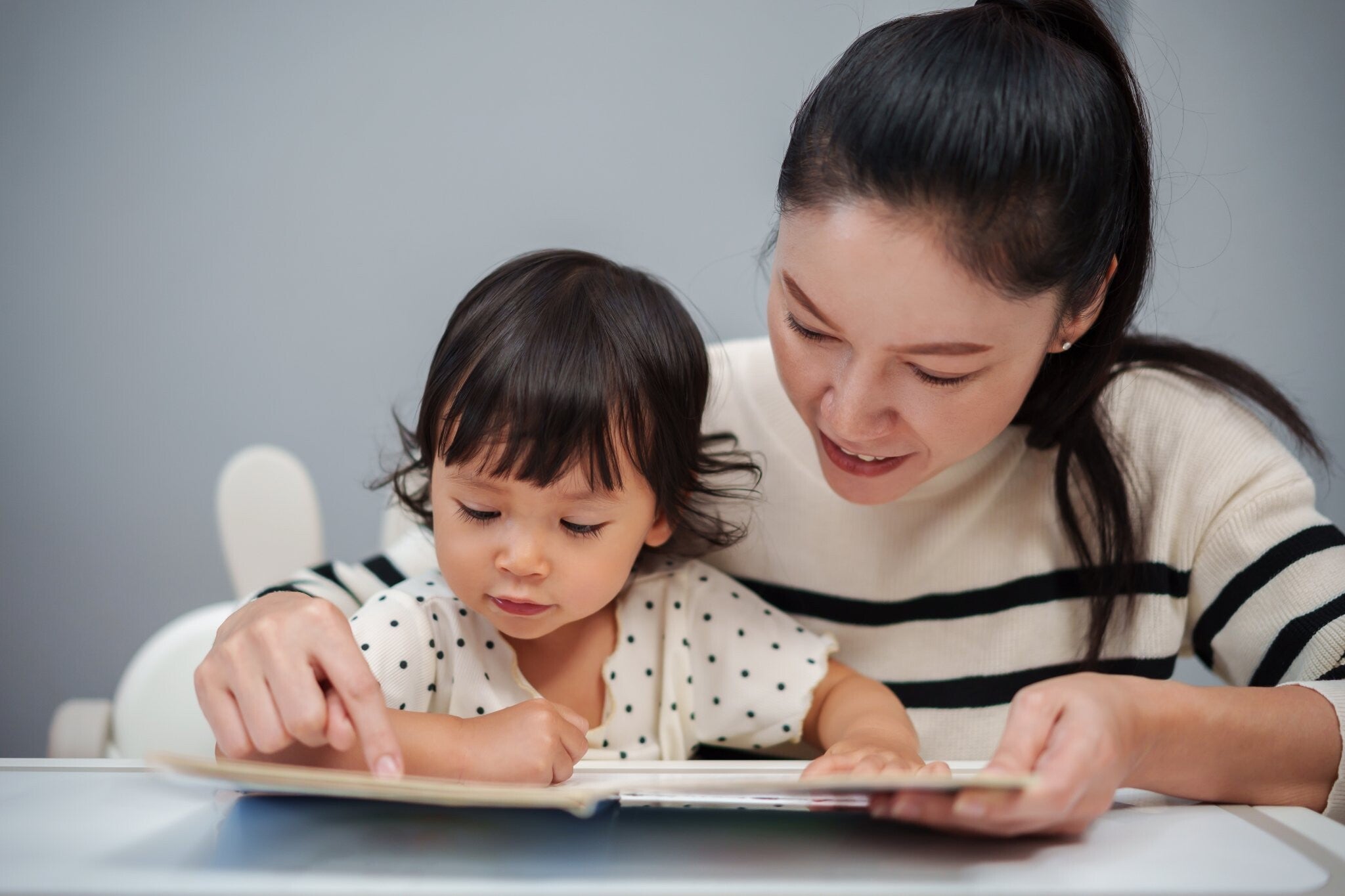 An Asian mom reads a Tagalog storybook to her toddler girl on the table.