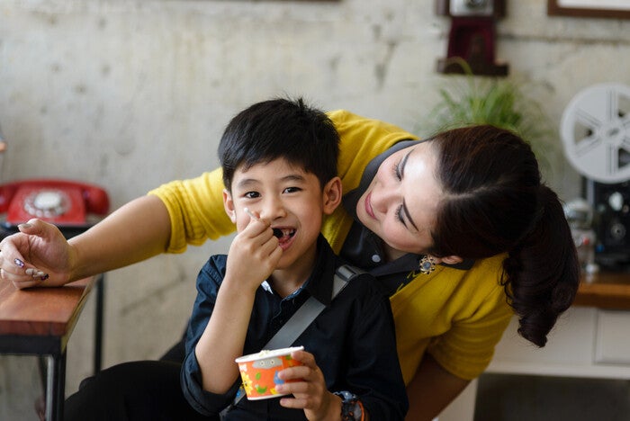 Asian single mom peeks at her son who’s eating ice cream during a day out, enjoying solo parent benefits.