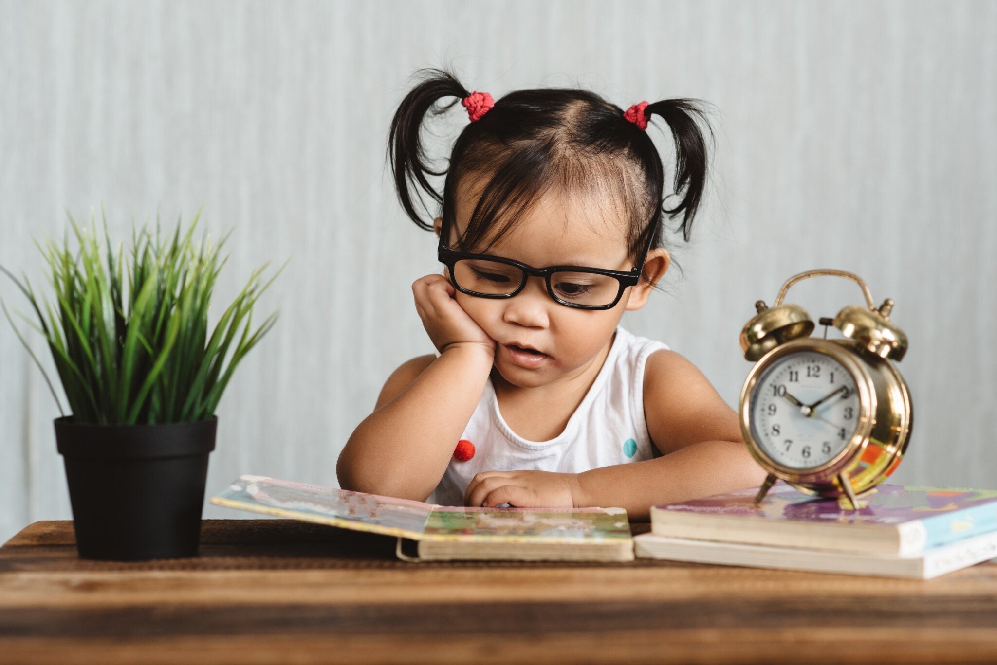A serious-looking Asian toddler girl wearing glasses and reading a book on a table with a clock and more books beside her.