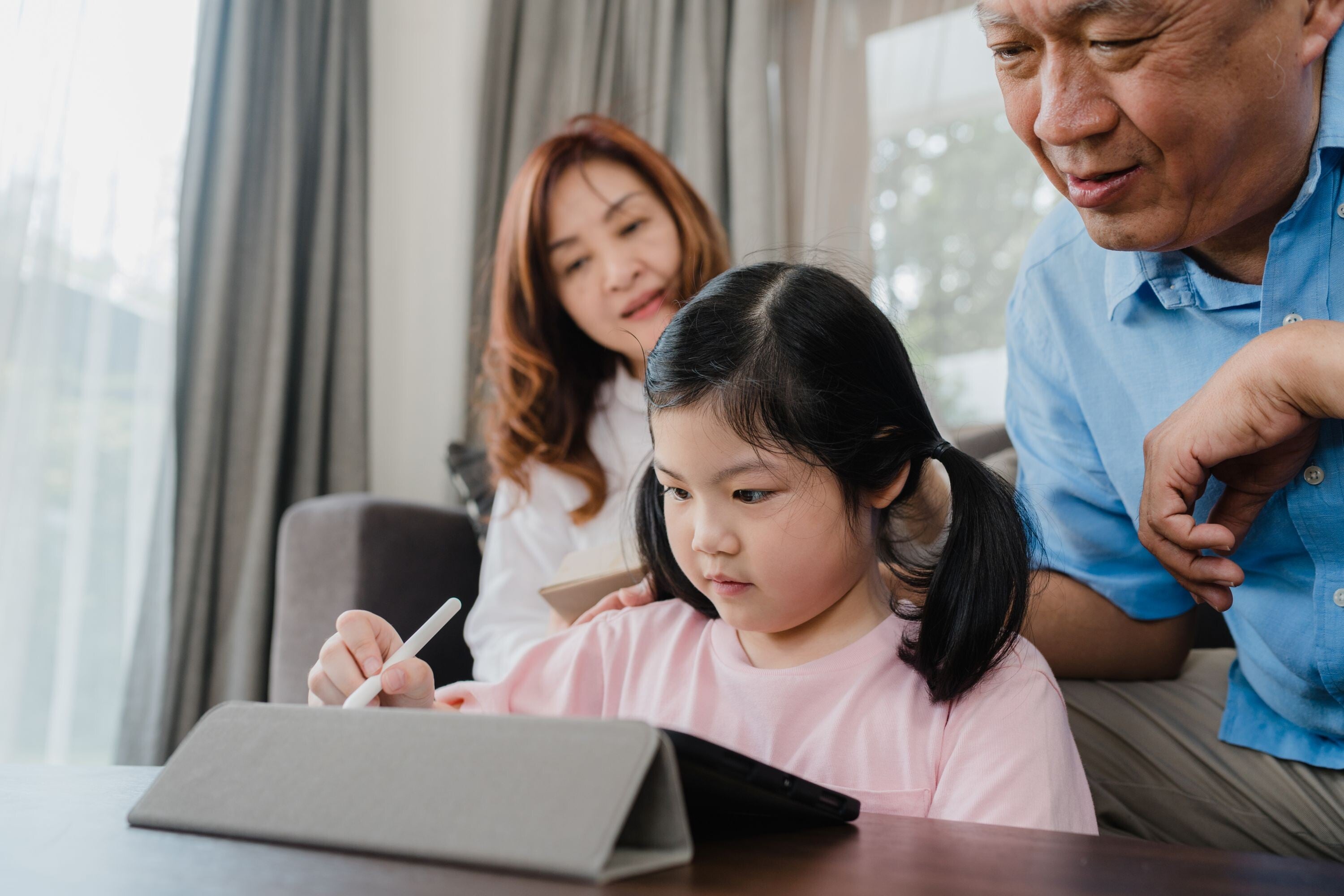 Asian woman and grandfather watching a little girl do her homework on an iPad.