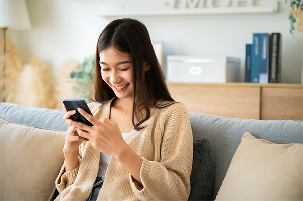 Happy Asian woman relaxing on a couch at home, texting messaging on smartphone.