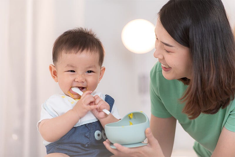 Asian baby boy bites into his spoon as his mom holds his bowl of food.