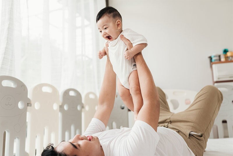 Asian dad lifts his healthy baby son while lying in bed
