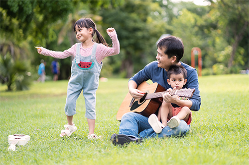 Asian dad playing the guitar while his daughter dances and his son sits on his lap in the park