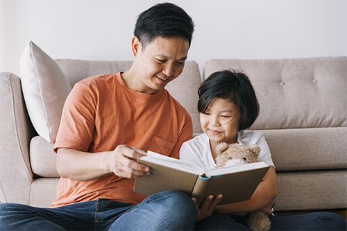 Asian father and daughter reading a book together by a sofa