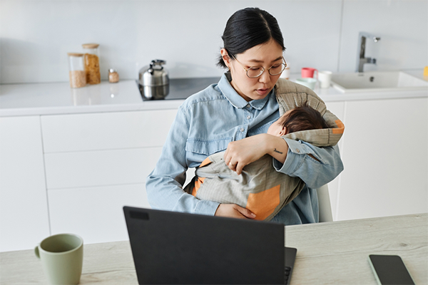 Asian mom adjusts her baby's position in a one-shouldered carrier as she works from home.