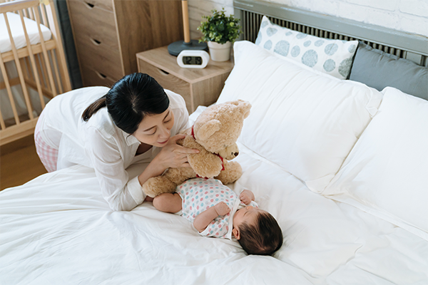 Asian mom hiding behind a teddy bear with her baby in bed, teaching object permanence, a baby milestone