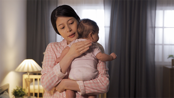 Asian mom in pajamas holding her newborn child by the bed at night.