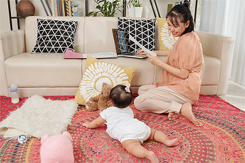 Asian mom looking at her baby crawling on the floor rug to get to a stuffed toy by the sofa