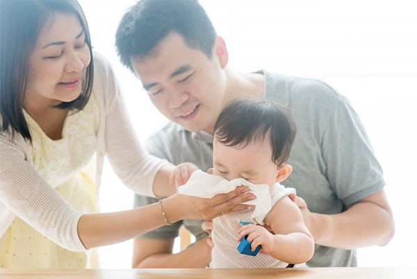 Asian mom wipes off baby's runny nose as he sits on his dad's lap